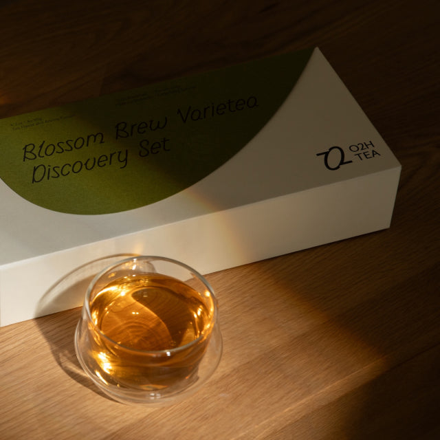 O2H Blossom Brew Varietea Discovery Set" elegantly presented beside a crystal clear glass of steeped black tea, capturing the essence of a premium tea tasting experience.